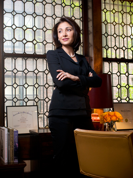 Professional portrait of a woman in a dark blazer standing with arms crossed in front of a geometric-patterned window, with a yellow armchair and orange floral arrangement nearby
